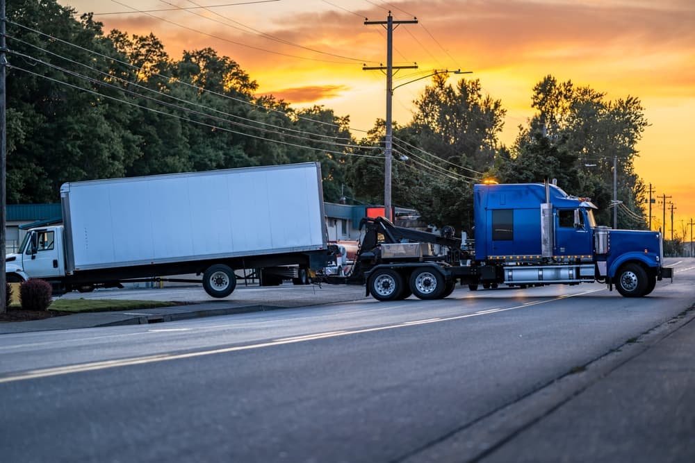 Mobile blue roadside assistance powerful tow big rig semi truck tractor towing broken after accident day cab semi truck with box trailer driving on the local road at evening time